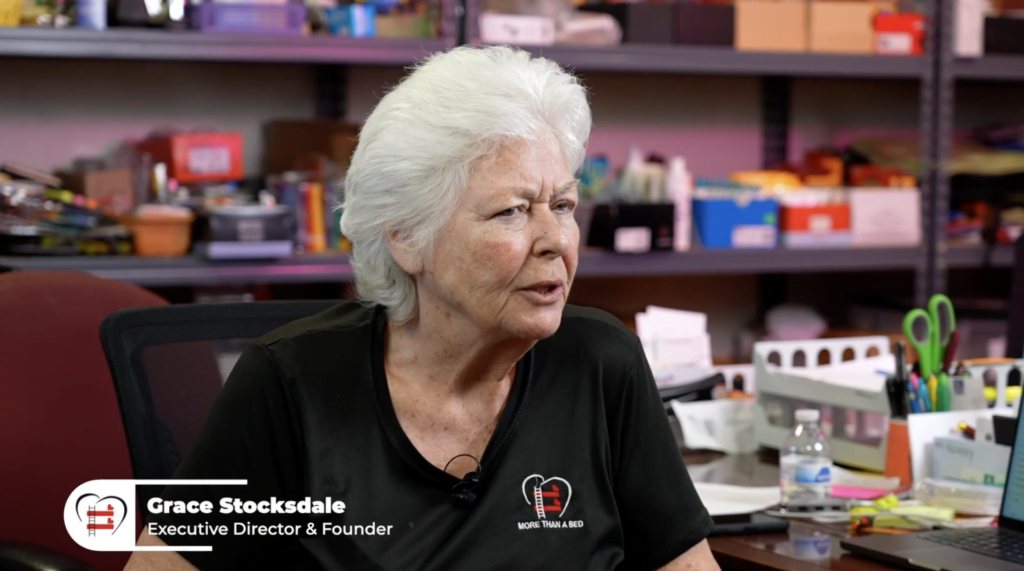 A woman in a More Than a Bed shirt speaks to the camera. Below her, text reads "Grace Stocksdale; Executive Director and Founder"